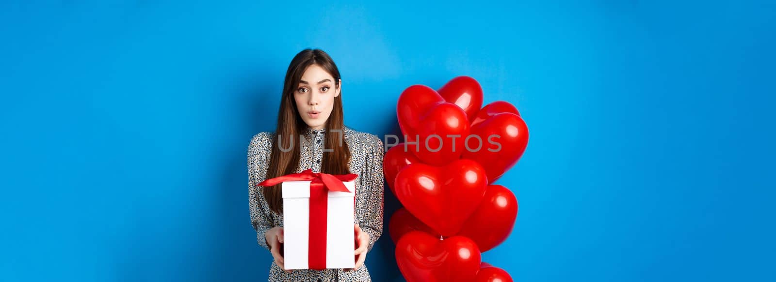 Valentines day. Surprised attractive girl looking amazed at camera, holding big romantic gift, standing near red hearts balloons, blue background by Benzoix