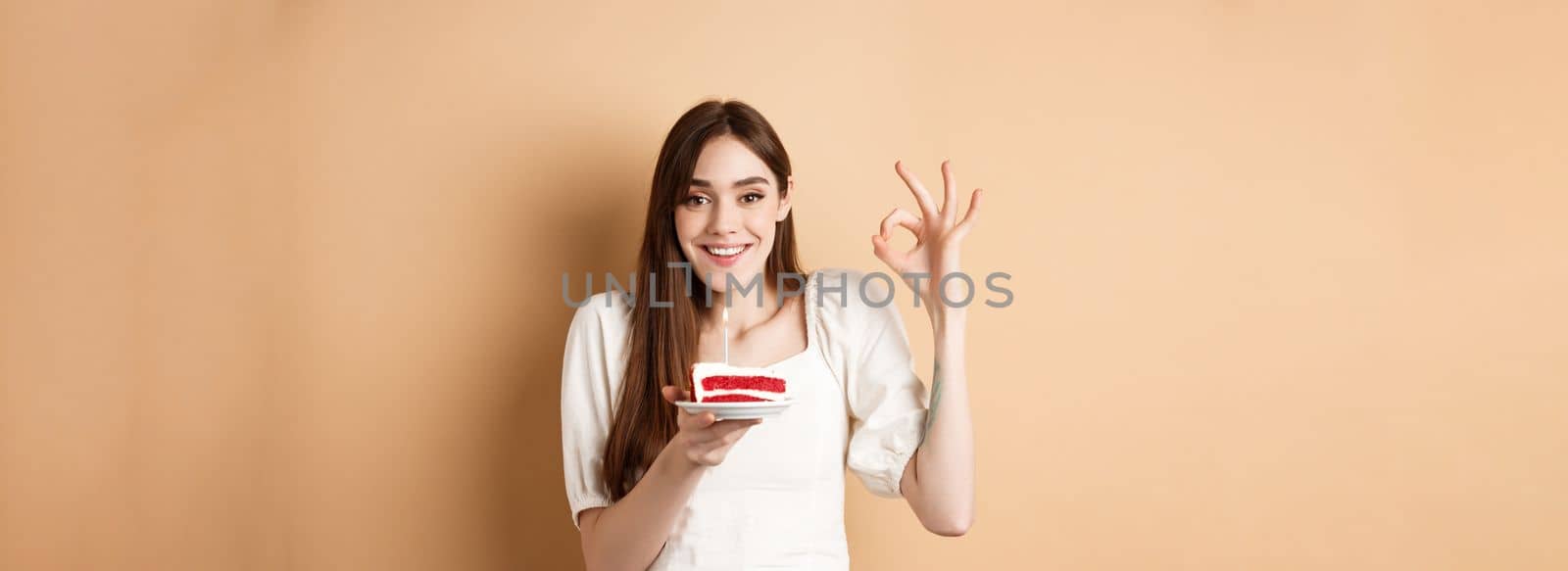 Happy birthday girl show okay gesture and hold bday cake, making wish on her holiday, standing on beige background.