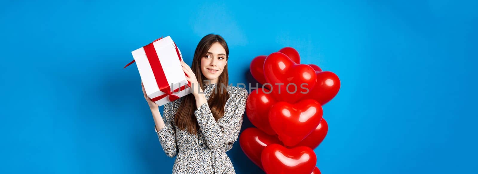 Valentines day. Beautiful woman shaking gift box to guess what inside, celebrating lovers holiday, standing near red hearts, blue background by Benzoix