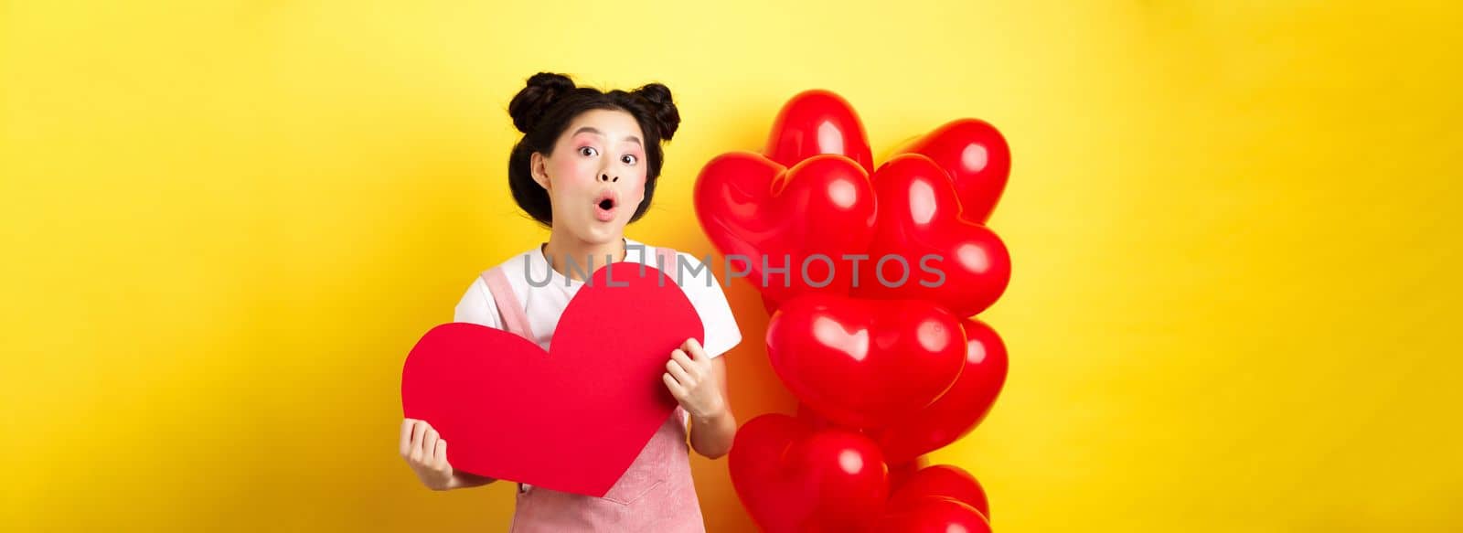 Happy Valentines day. Silly asian girl showing big red heart postcard and looking amazed, standing near red romantic balloons, yellow background.