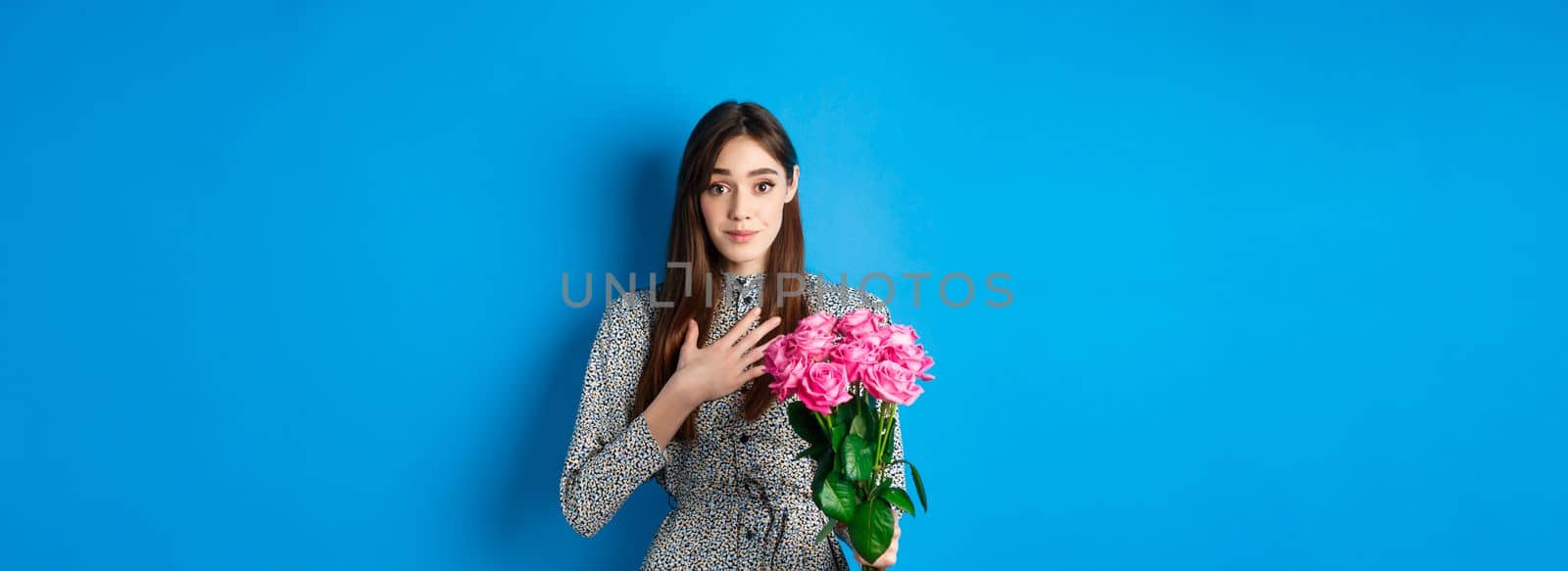 Valentines day concept. Touched and thankful pretty girl, holding hand on heart and smiling grateful for gift, holding bouquet of roses, standing on blue background by Benzoix