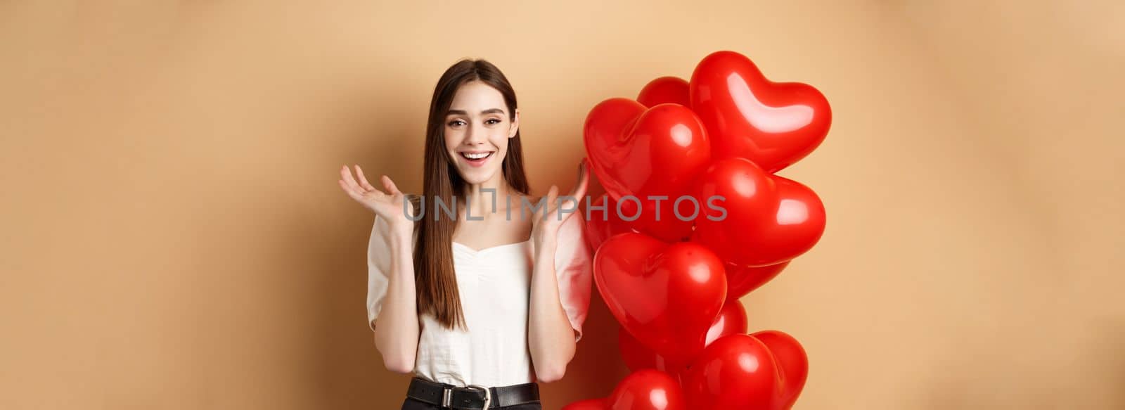 Valentines day and love concept. Happy smiling woman raising hands up surprised, standing near romantic heart balloons on beige background.
