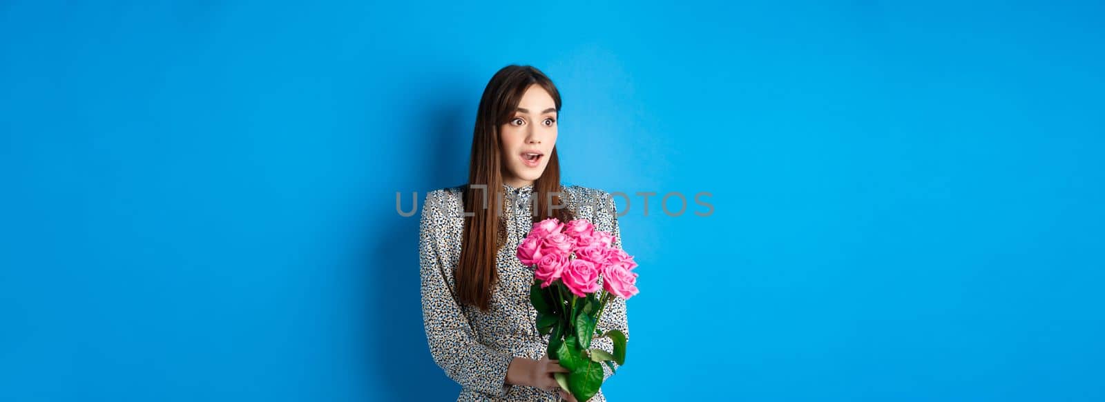 Valentines day concept. Surprised happy girl looking aside with amazement, receiving bouquet of romantic flowers, holding pink roses, standing in dress on blue background by Benzoix