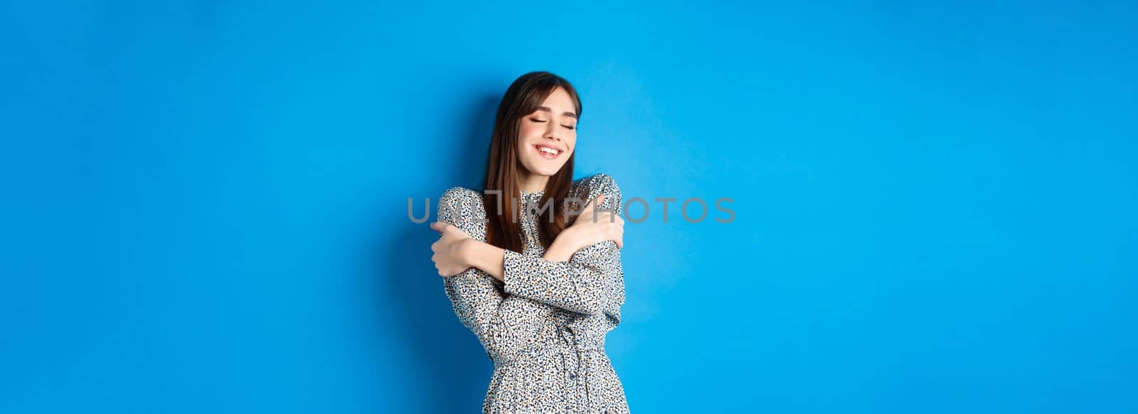 Smiling romantic girl in dress hugging herself, close eyes and feel happy, standing on blue background by Benzoix