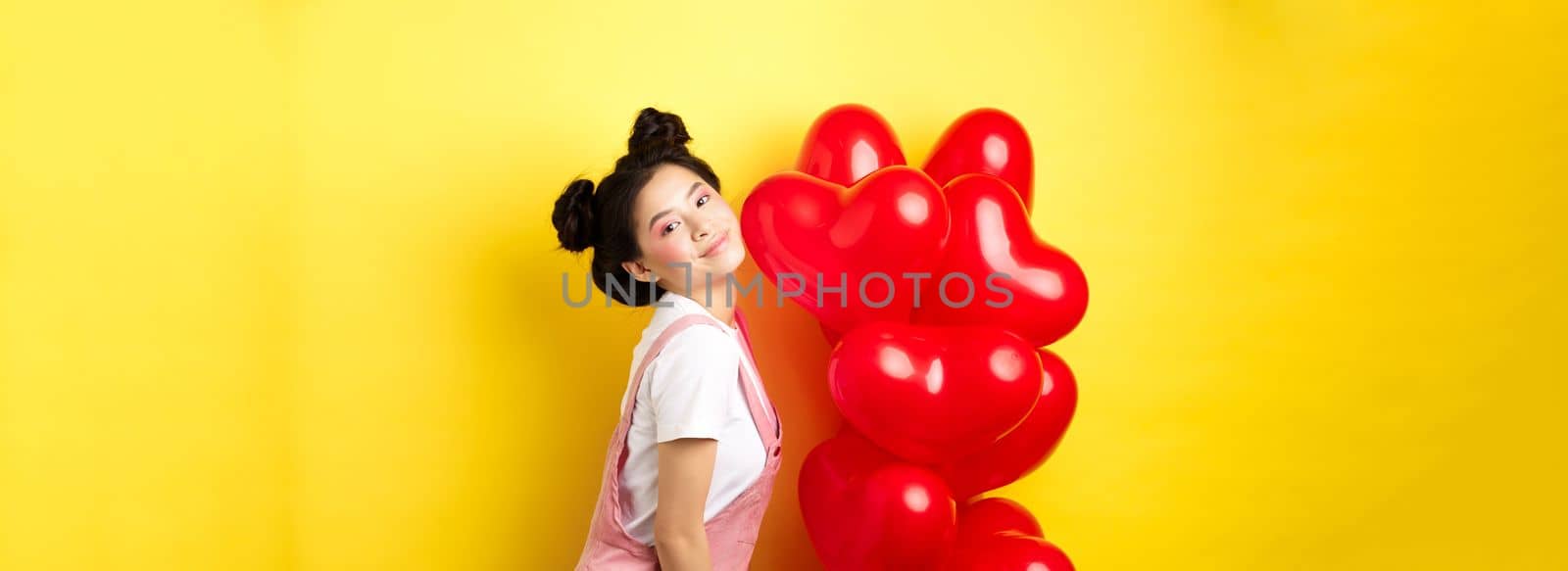 Valentines day concept. Stylish teenage asian girl posing near red hearts balloons, wear outfit for romantic date, standing happy on yellow background.