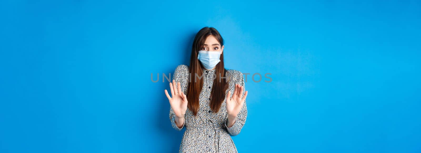 Covid-19, pandemic lifestyle concept. No thank you. Reluctant and worried girl in medical mask asking non-masker to stay away, showing block gesture, tell stop, blue background by Benzoix