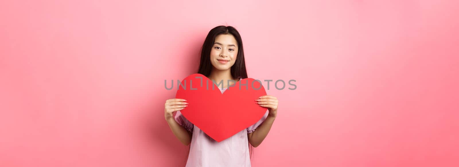 Valentines day concept. Cute teenage asian girl showing big red heart card, falling in love, going on romantic date in dress, smiling tender at camera, pink background by Benzoix