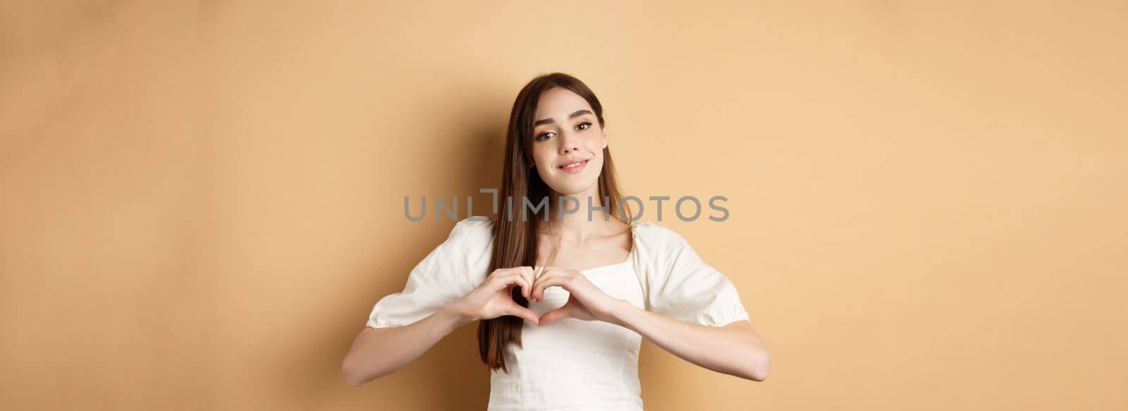 Beautiful caucasian woman say I love you, showing heart gesture and smile at camera, beige background. Concept of romance and Valentines day by Benzoix
