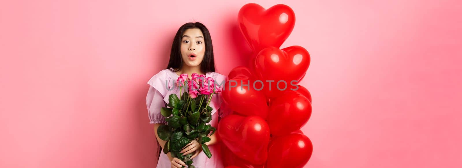 Surprised asian girl in dress standing near valentines day heart balloons and say wow at camera, holding flowers bouquet from lover, romantic date with roses, pink background.