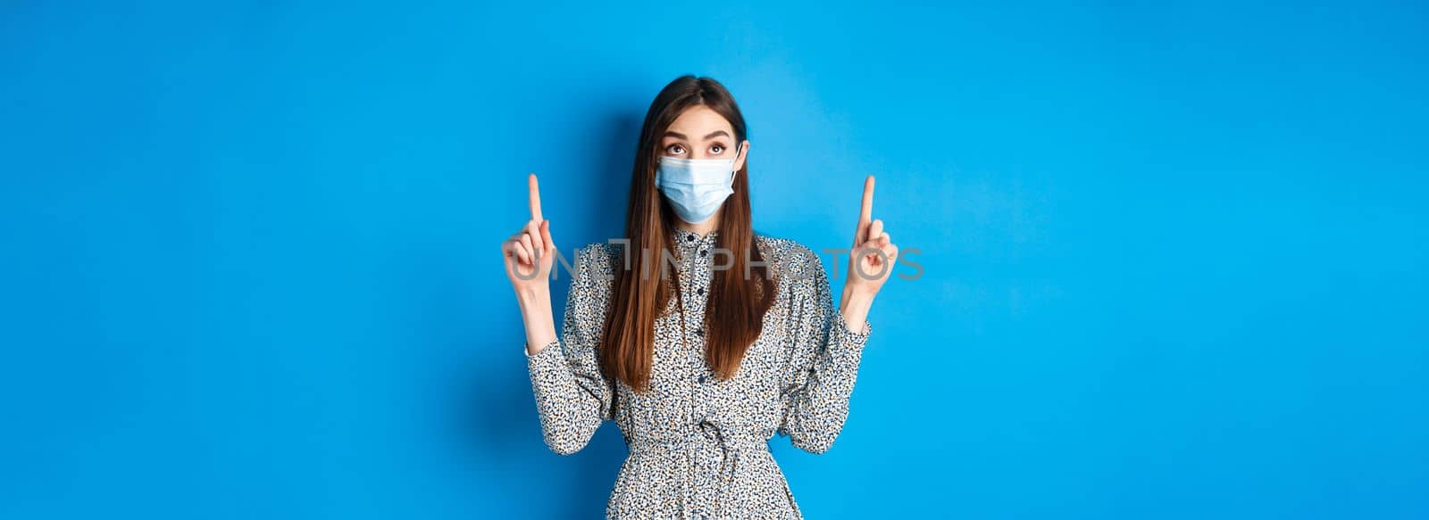 People, covid and quarantine concept. Intrigued young woman looking and pointing up at advertisement, standing in dress and face mask from coronavirus, blue background.