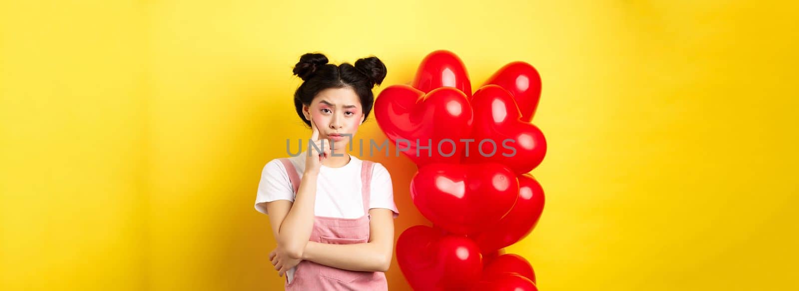 Valentines day concept. Unamused and bored asian girlfriend staring skeptical at camera, waiting for something near red hearts balloons, yellow background by Benzoix