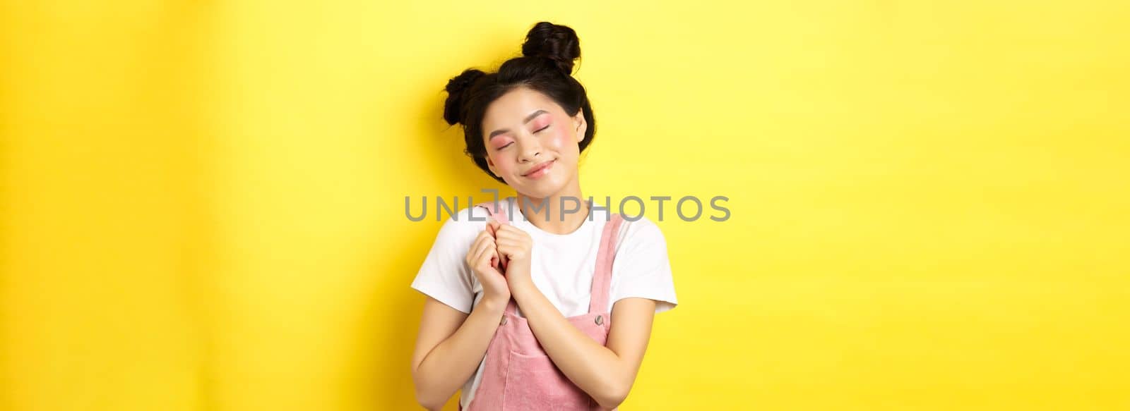 Cute asian girl with makeup, close eyes and remember beautiful moment, holding hands on heart daydreaming, standing on yellow background by Benzoix