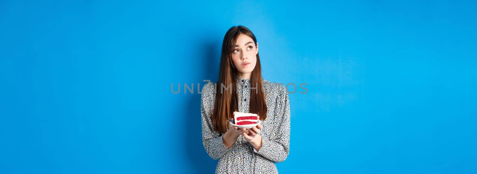 Birthday girl making wish, looking aside at logo and holding b-day cake, standing on blue background by Benzoix
