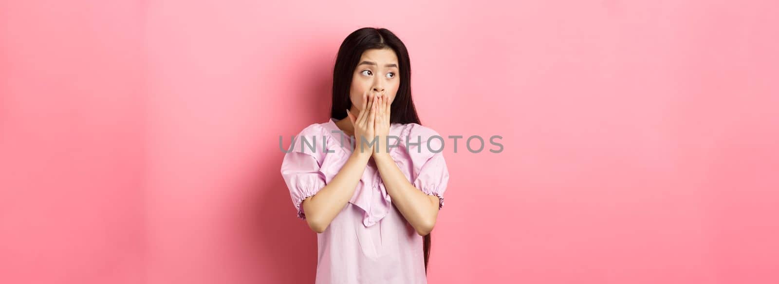 Shocked and worried asian woman looking aside at empty space with concerned face, covering mouth with hands, standing in dress on pink background.