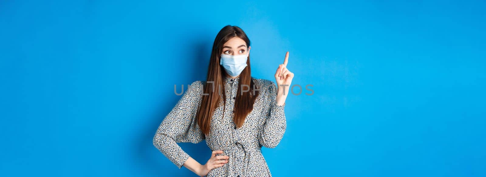 People, covid and quarantine concept. Excited cute female in medical mask, pointing and looking at logo with happy amazed face, standing on blue background.