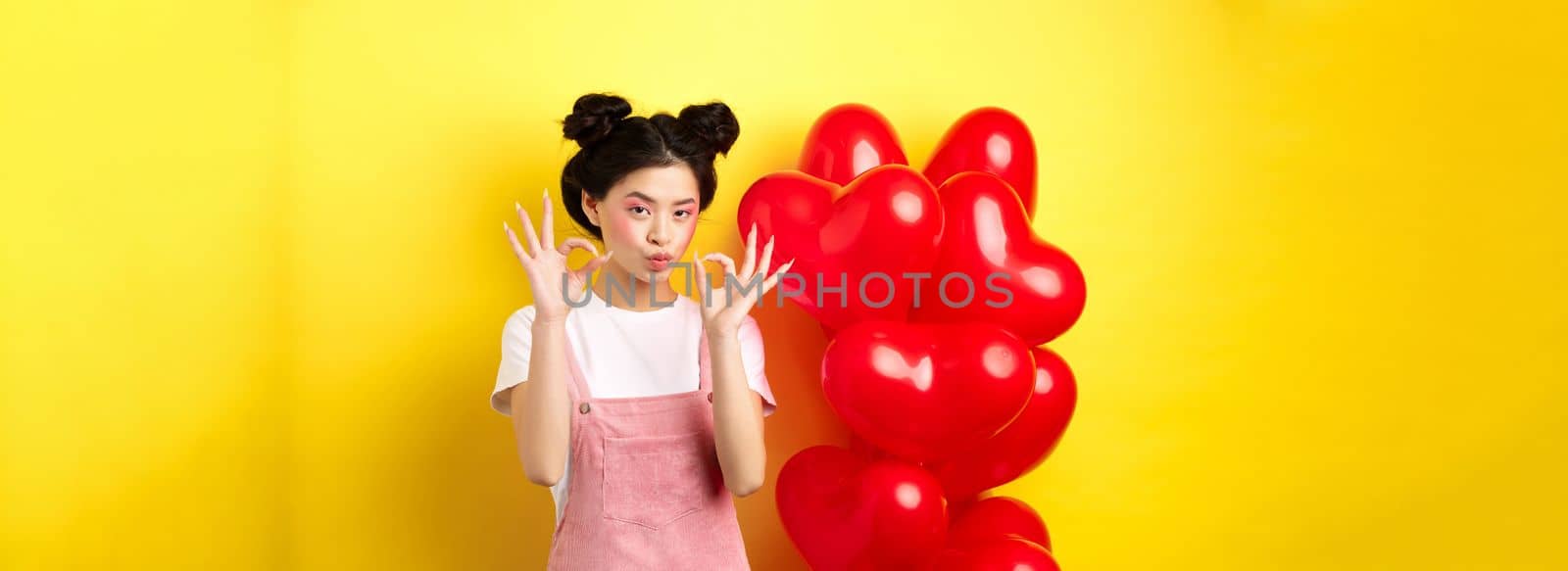Cute young asian woman in stylish clothes and makeup, showing okay signs near st Valentines day heart balloons, praise good offer, standing on yellow background.