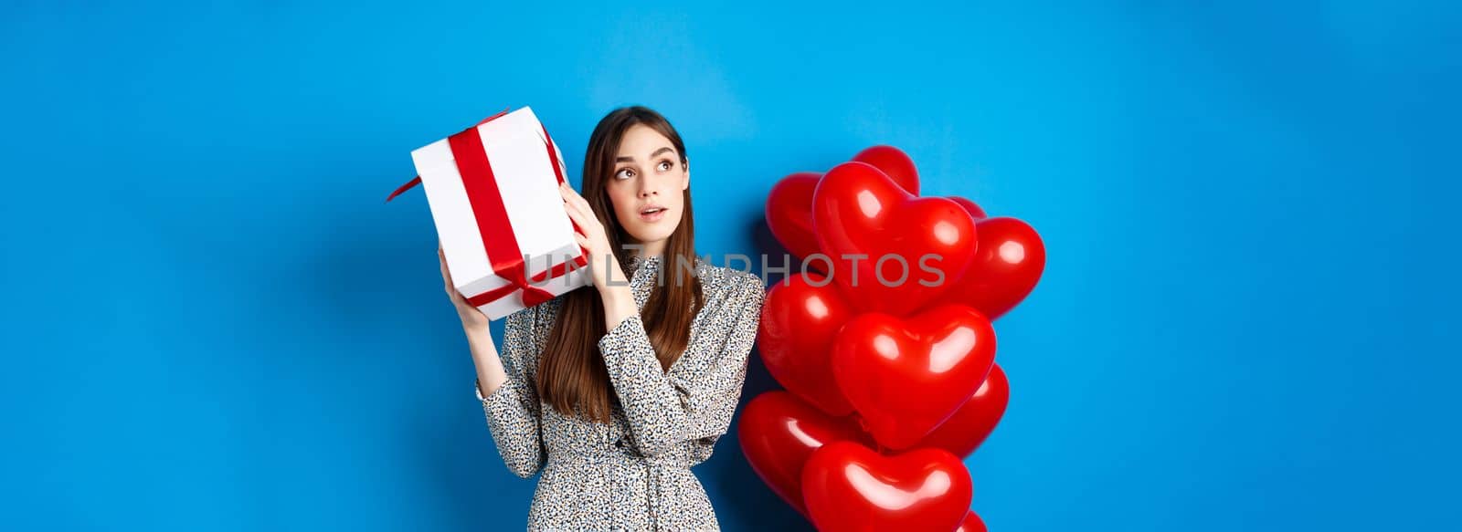 Valentines day. Beautiful woman shaking gift box to guess what inside, look dreamy, celebrating lovers holiday, standing near red hearts, blue background.