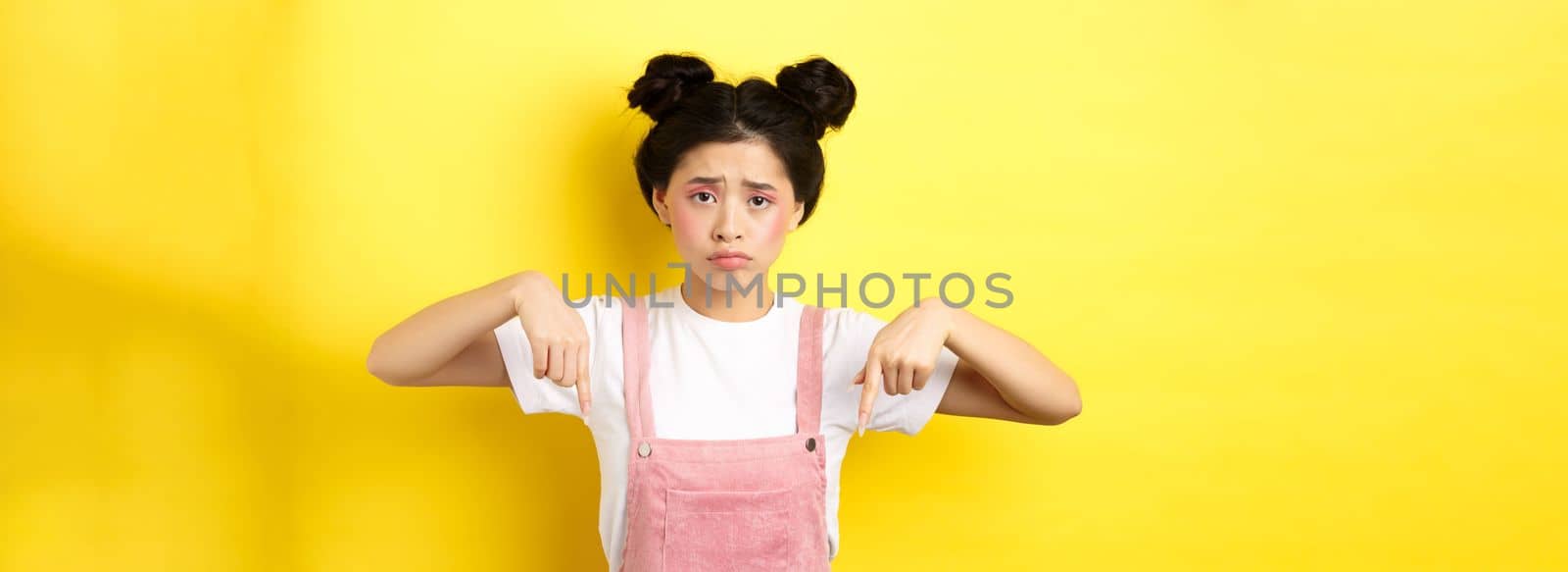 Sad frowning asian girl pointing fingers down at unfair thing, looking upset and gloomy, standing with makeup on yellow background.