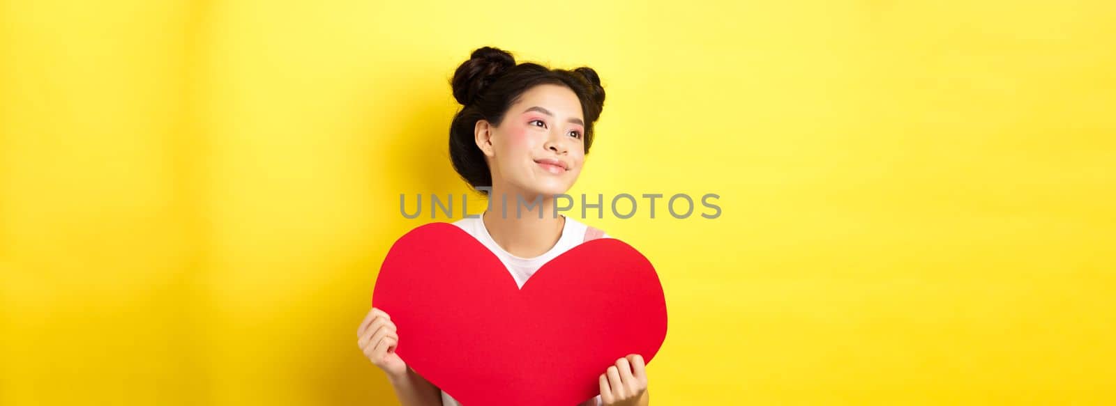 Asian girl in love, looking dreamy left and smiling, showing Valentines red heart cutout, imaging romantic date, standing on yellow background.