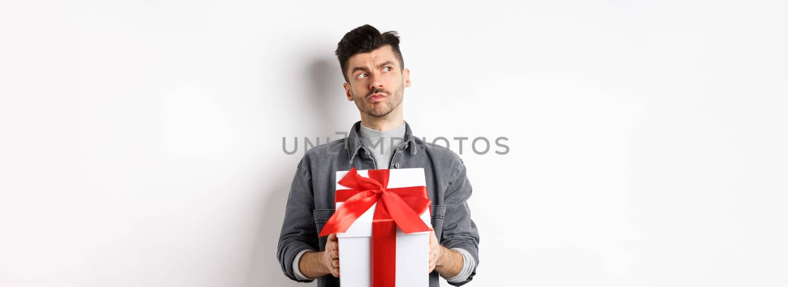 Pensive boyfriend looking aside and holding gift box, waiting for lover, making surprise present on valentines day, planning date with girlfriend, white background.