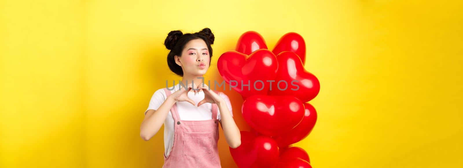 Beautiful asian woman showing heart, I love you gesture and kissing lips, standing near romantic red balloons. Concept of Valentines day.