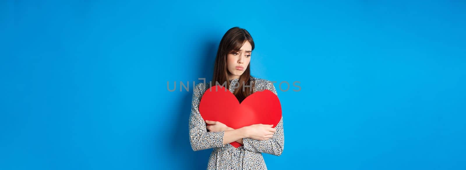 Valentines day. Lonely single girl hugging big red heart cutout and looking down upset, standing gloomy on blue background.