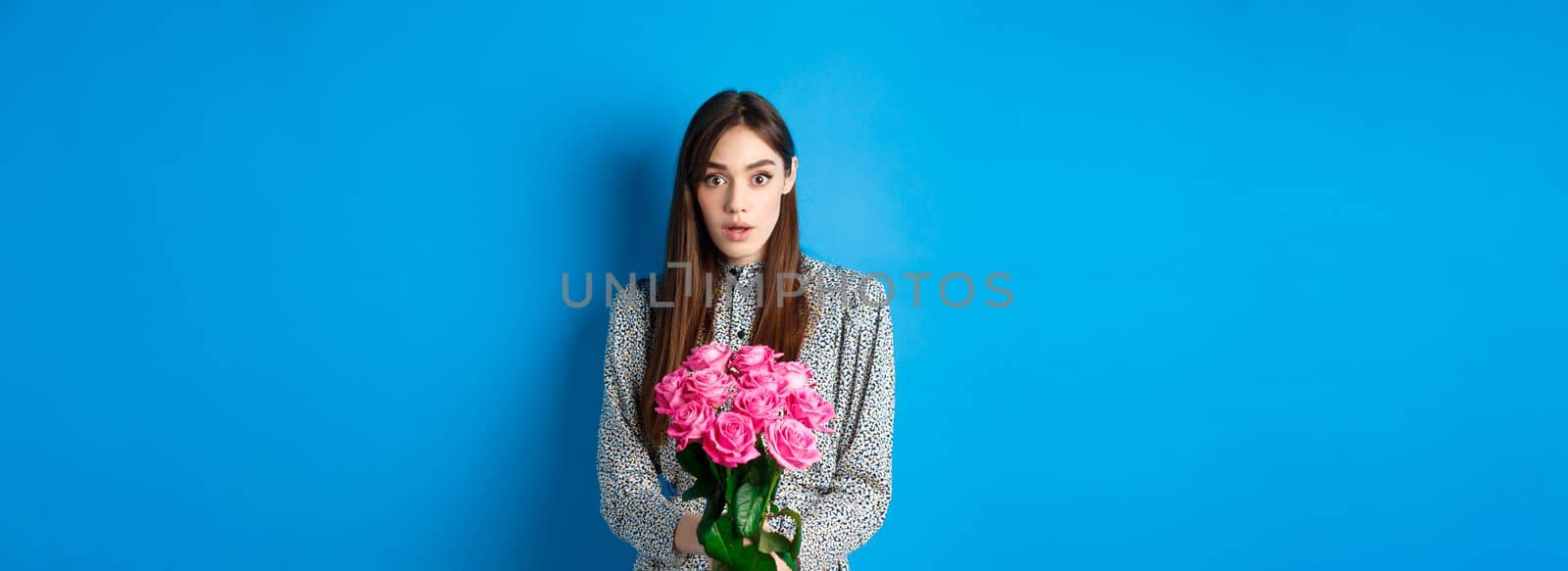 Valentines day concept. Surprised girlfriend receiving beautiful bouquet of flowers and looking with disbelief at camera, standing on blue background.