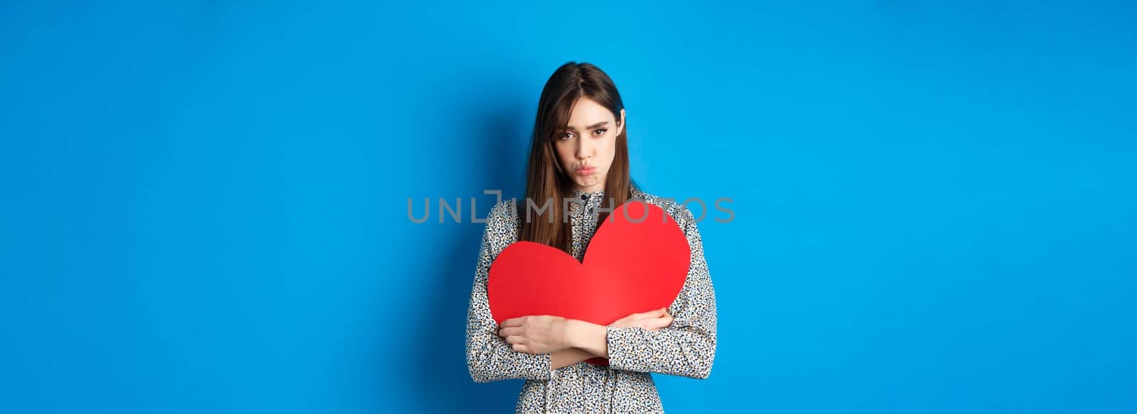 Valentines day. Sad and gloomy caucasian girl pucker lips and looking disappointed, holding big red heart cutout, standing on blue background by Benzoix