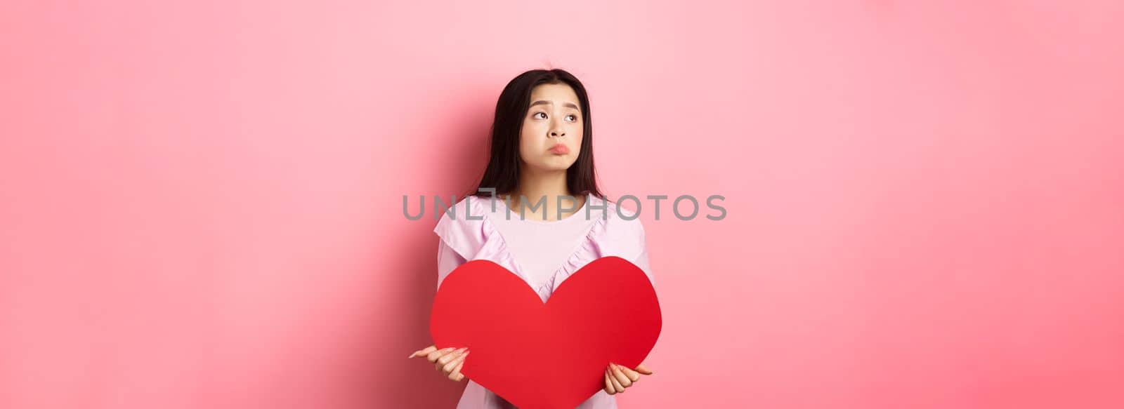 Valentines concept. Lonely teenage asian girl dreaming about love, feeling sad and lonely on lovers day, looking aside with pity, holding big red heart, pink background.