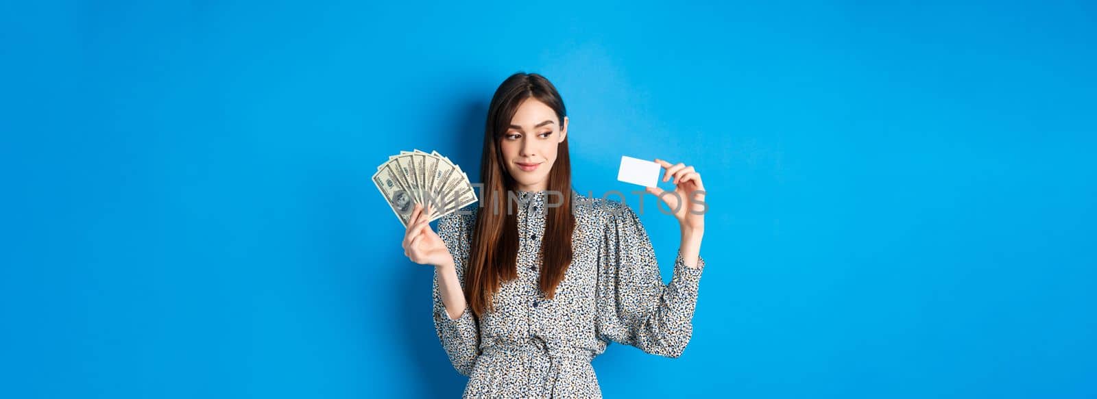 Shopping. Beauty girl in dress looking aside and smiling pensive, showing money and plastic credit card, standing on blue background.