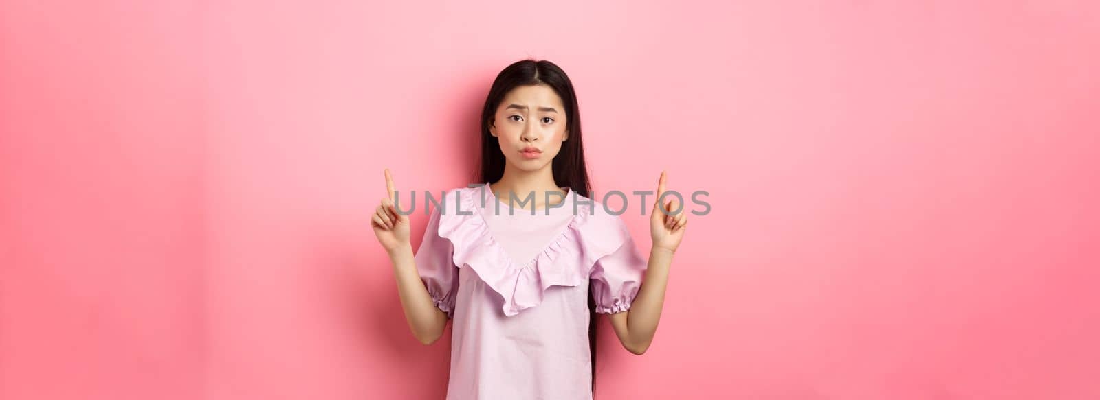 Cute timid asian woman pointing fingers up, frowning and look upset, pointing fingers up at logo, standing in dress on pink background by Benzoix