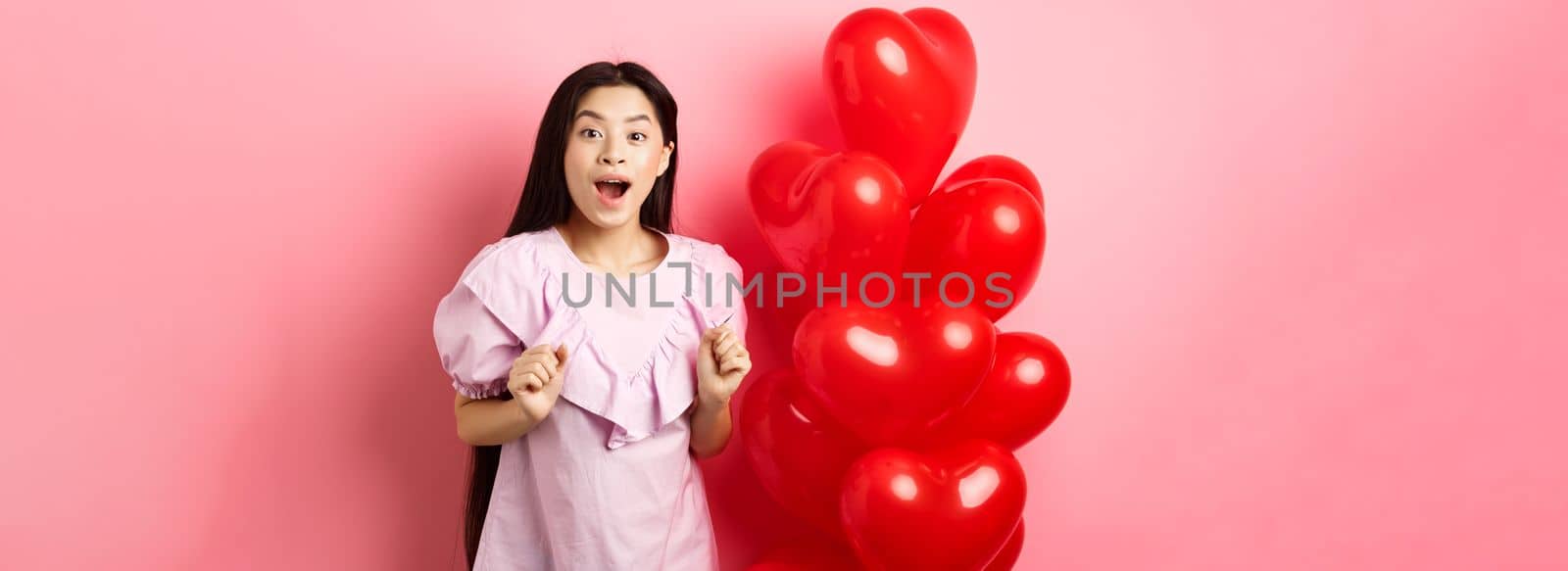 Excited and surprised teenage korean girl open mouth amazed, receive surprise gift on valentines day, looking wondered, standing near heart balloons, pink background by Benzoix