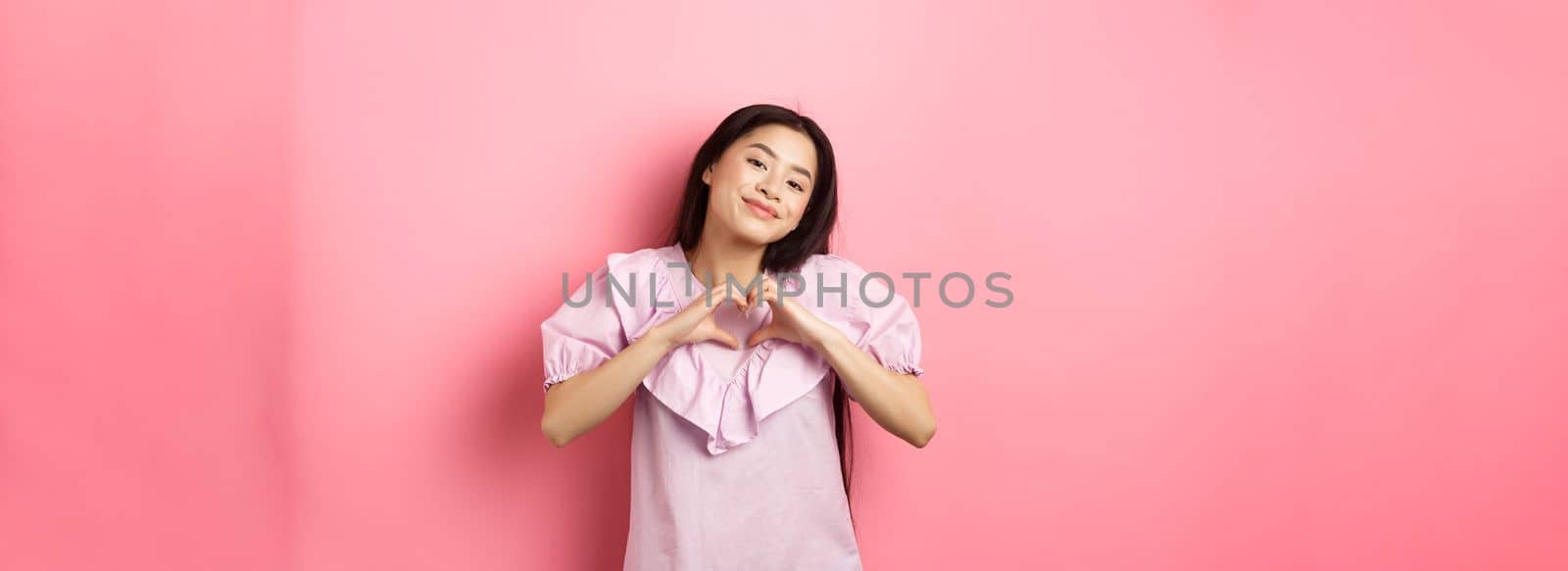 Valentines day concept. Beautiful korean teenager in dress showing heart gesture and smiling with admiration and affection, falling in love, feel romantic, pink background by Benzoix