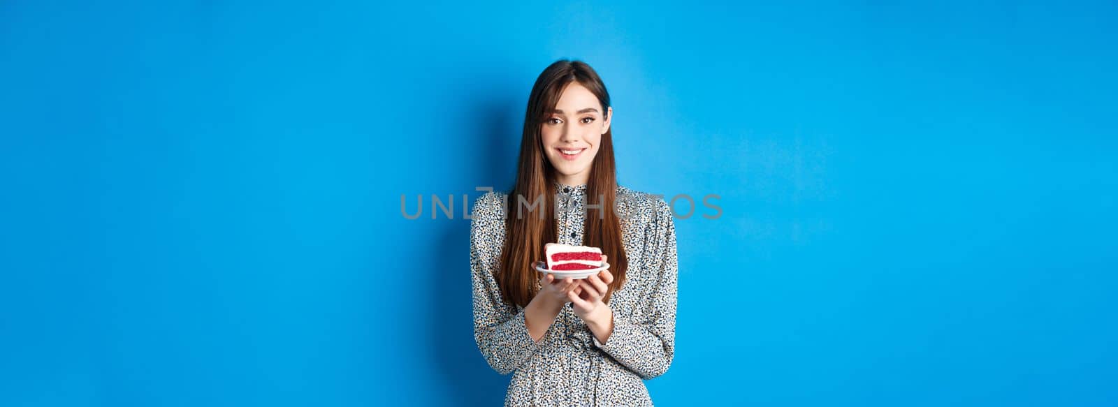 Beautiful girl holding birthday cake and celebrating, wishing happy bday and smiling, standing against blue background.