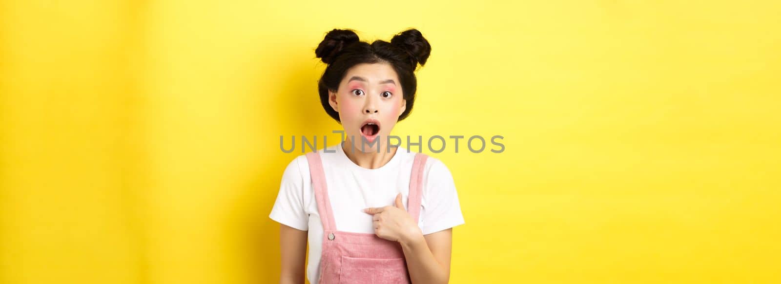 Surprised asian teen girl with glamour makeup, pointing at herself and gasping confused, being chosen or accused, standing on pink background.