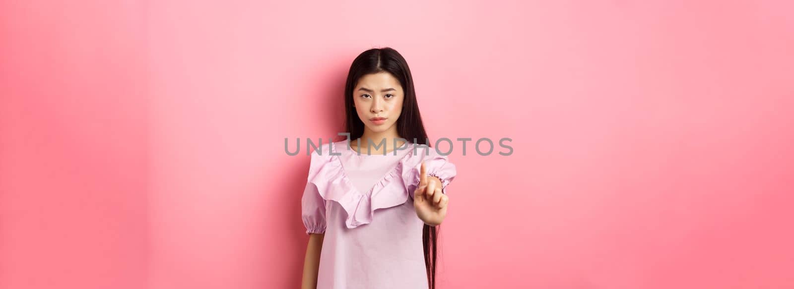 She says no. Serious asian girl shaking finger in stop gesture, prohibit and disagree with person, scolding bad behaviour, standing against pink background.