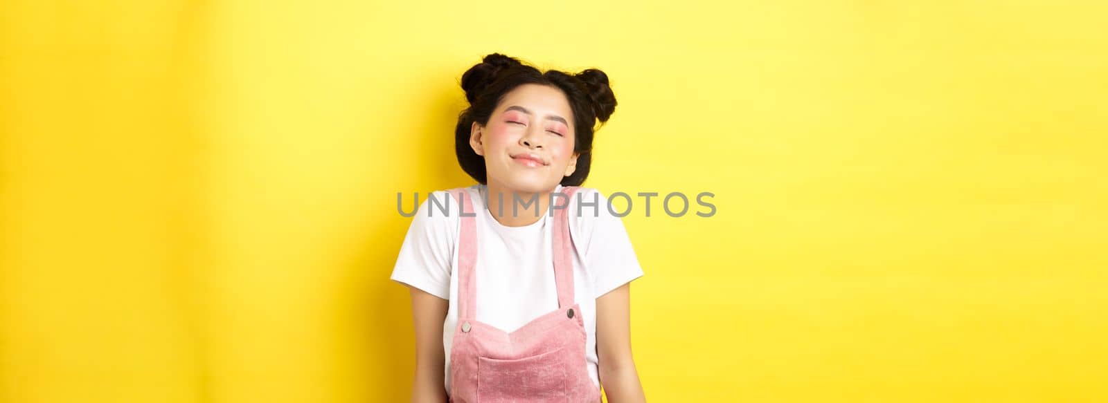 Romantic asian girl with cute pink makeup and summer clothes, close eyes and smiling dreamy, standing on yellow background.