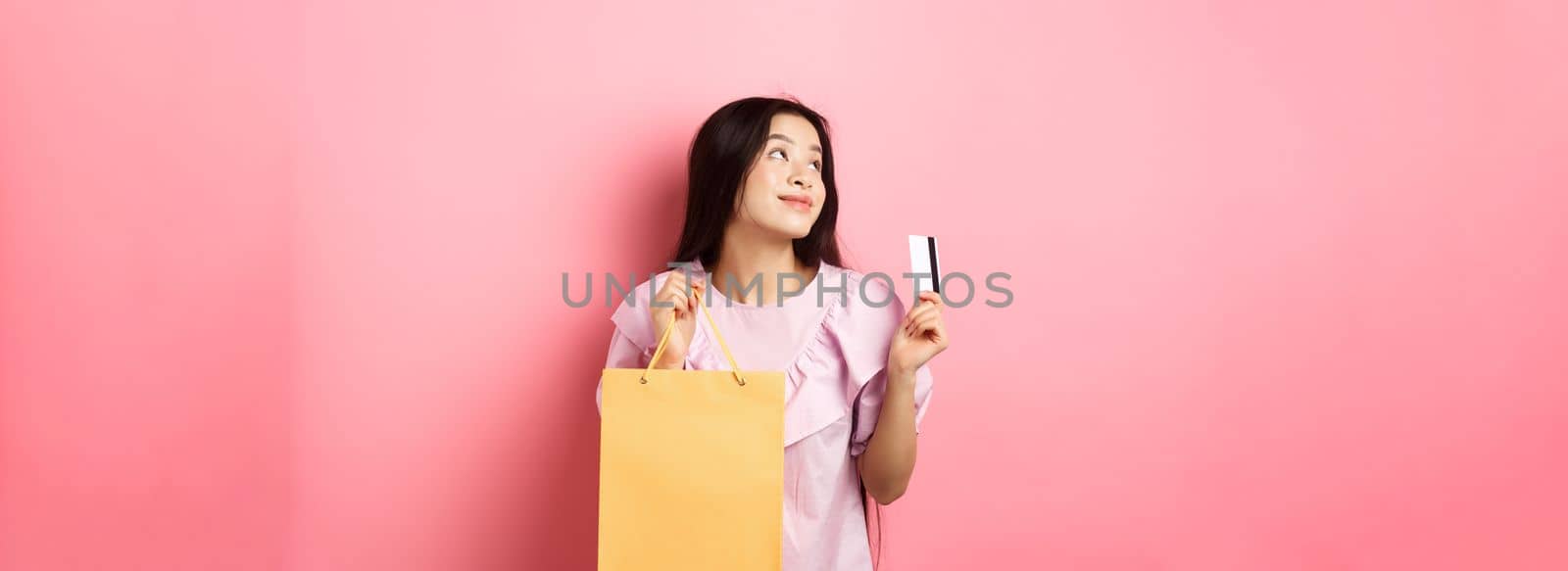 Shopping. Dreamy asian girl thinking of buying something, holding shop bag and plastic credit card, standing against pink background by Benzoix