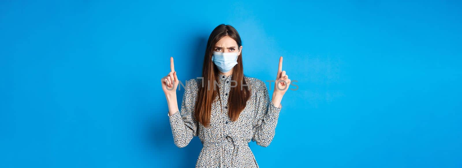 Covid-19, social distancing and healthcare concept. Angry woman frowning in medical mask, pointing and looking up with condemn and disappointment, blue background.