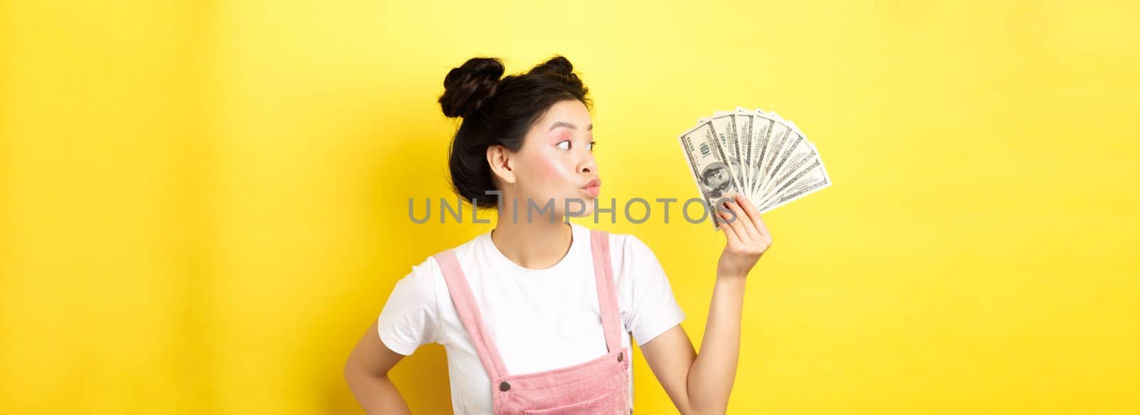 Shopping. Silly korean girl with glam makeup, pucker lips and looking at dollar bills, wasting money, standing on yellow background.