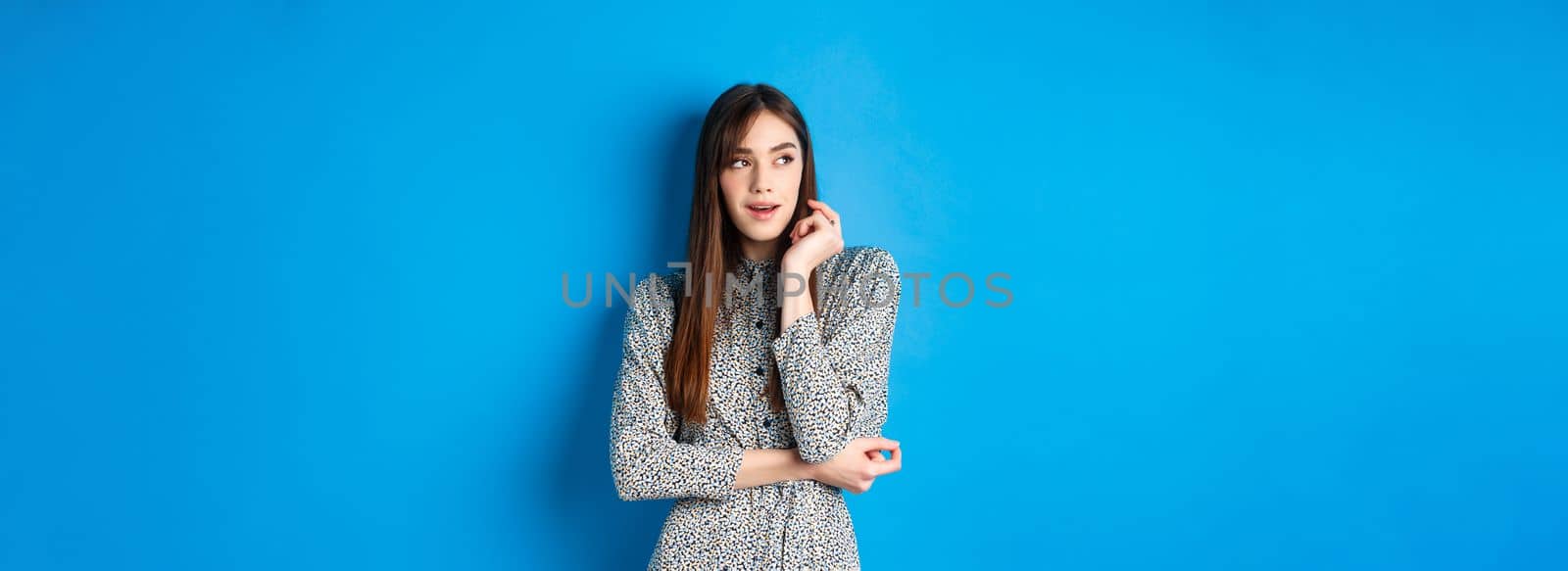 Pensive caucasian girl with long natural hair, wearing vintage dress, looking left with thoughtful face, standing on blue background by Benzoix