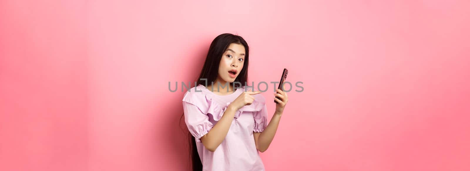 Excited beautiful asian woman, pointing finger at smartphone and look curious, checking out online promo, standing against pink background.