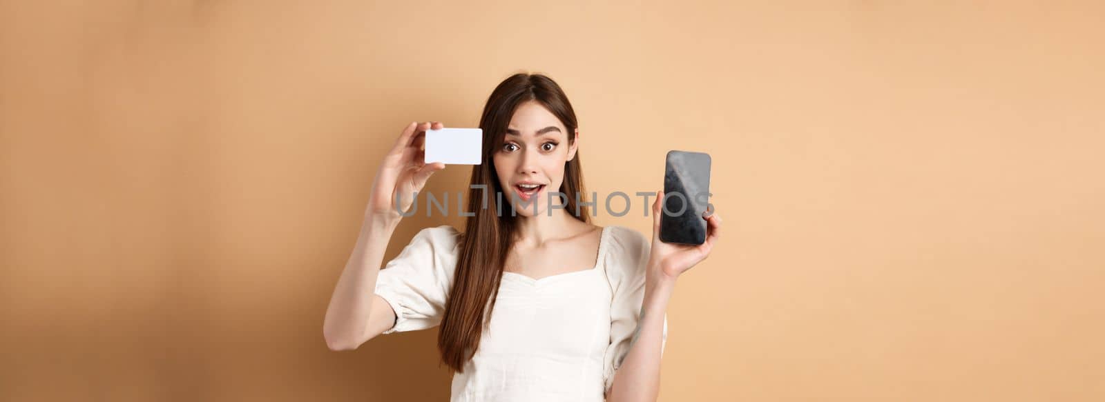Excited girl showing plastic credit card of bank and empty mobile phone screen, demonstrate shopping app, standing on beige background by Benzoix
