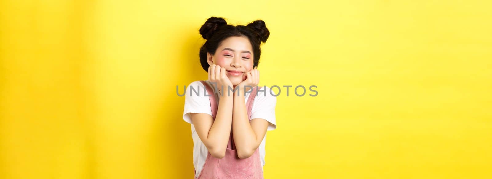 Cute happy asian woman with bright makeup and dungarees, leaning satisfied on hands and smiling, standing on yellow background by Benzoix