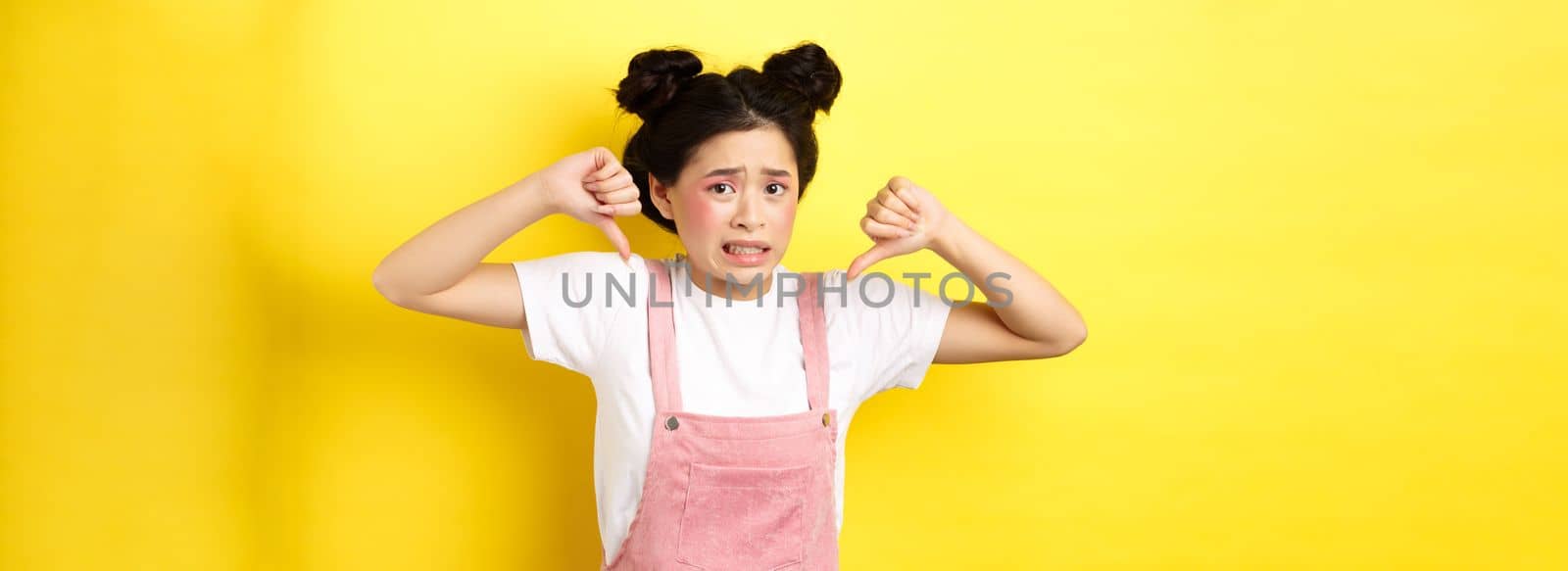 Disgusted asian girl showing thumbs down and cringe from bad product, standing in summer clothes against pink background by Benzoix