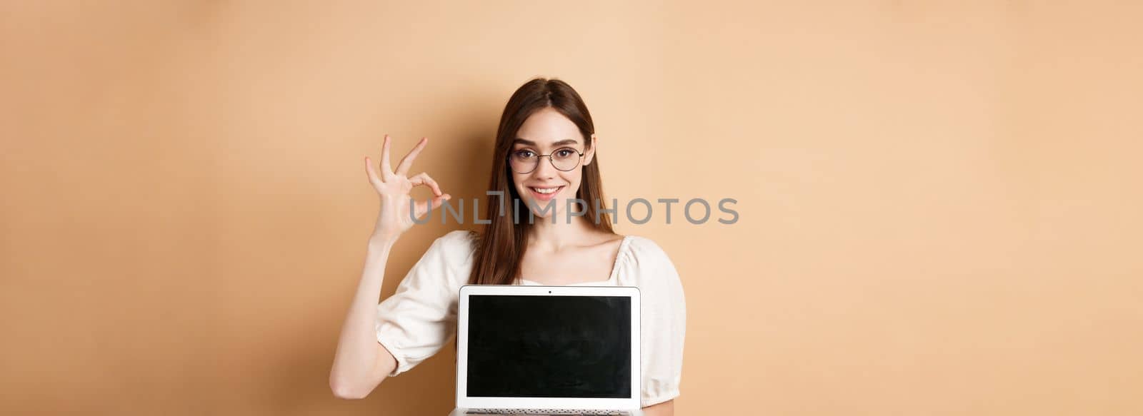 E-commerce. Smiling young woman in glasses showing okay sign and laptop screen, recommending internet promo, standing on beige background.