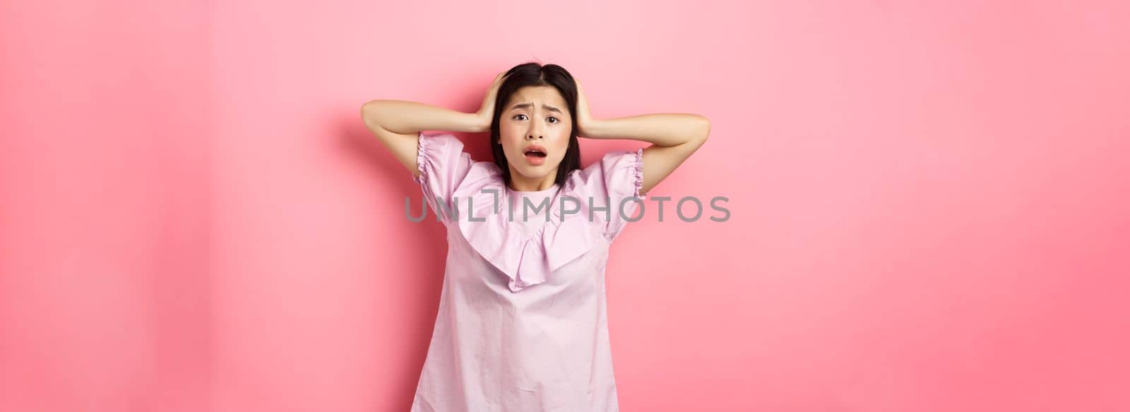 Frustrated asian teen girl panicking, holding hands on head and screaming scared, standing anxious on pink background.