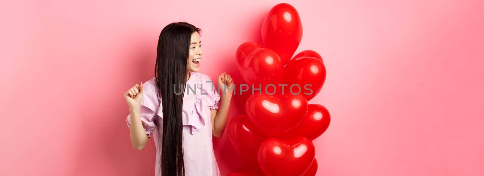 Asian teenage girl with long hair, cheering from valentines day romantic gift, looking at logo and smiling happy, jumping from joy near lovers gift heart balloons, pink background.