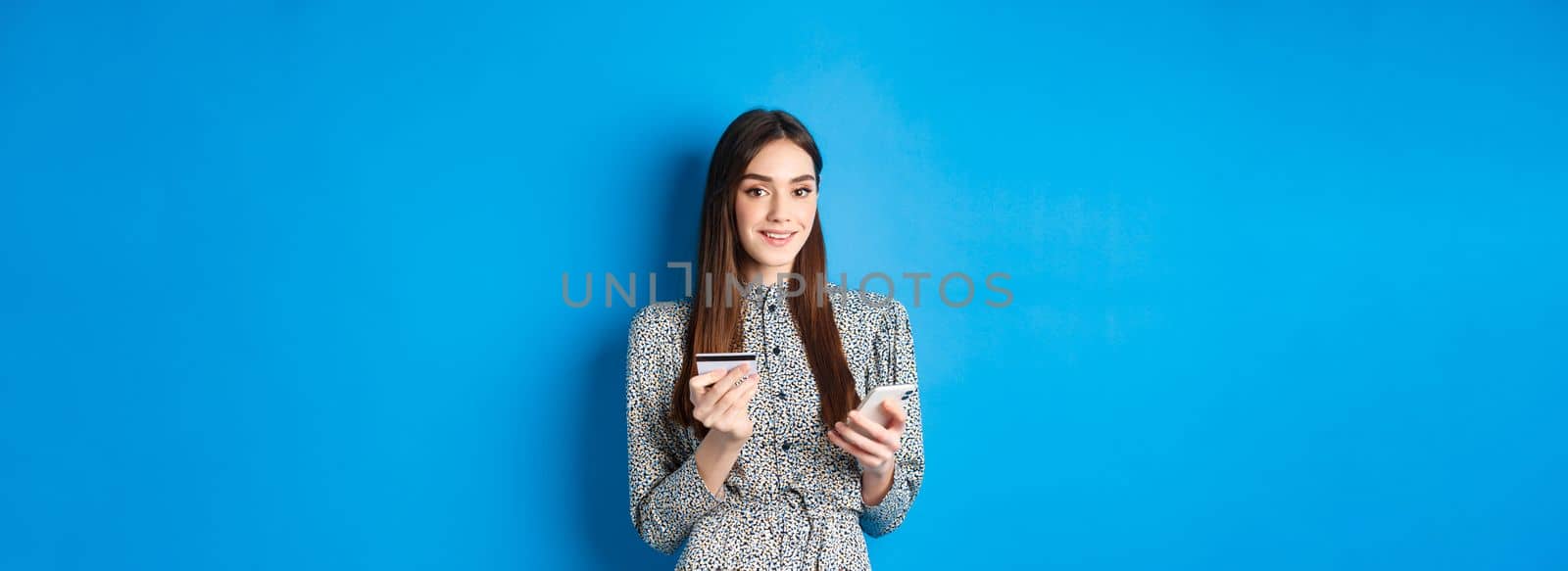 Online shopping. Happy smiling woman buying in app, holding smartphone and plastic credit card, blue background.