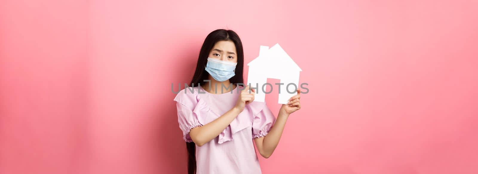 Real estate and pandemic concept. Gloomy girl showing paper house cutout, wearing medical mask and dress, standing against pink background.